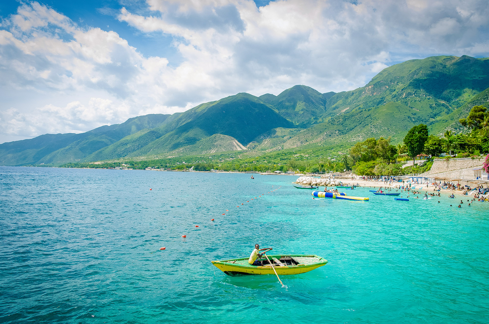 Boats and people swimming on the beach at Wahoo Bay, Cotes des Arcadins, Haiti, with mountains in the background