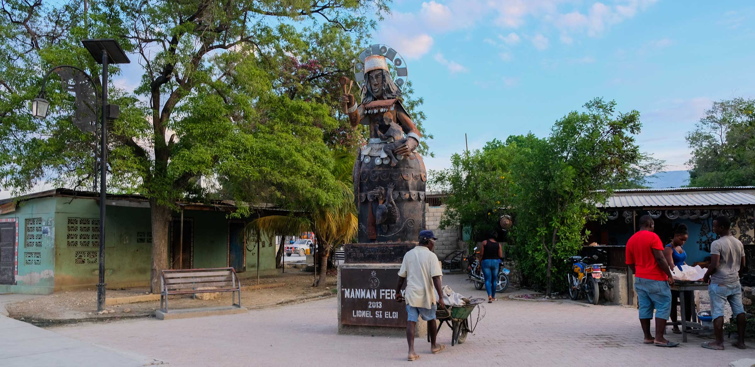 Man walks with wheelbarrow past metal statue in Village Noailles, Haiti