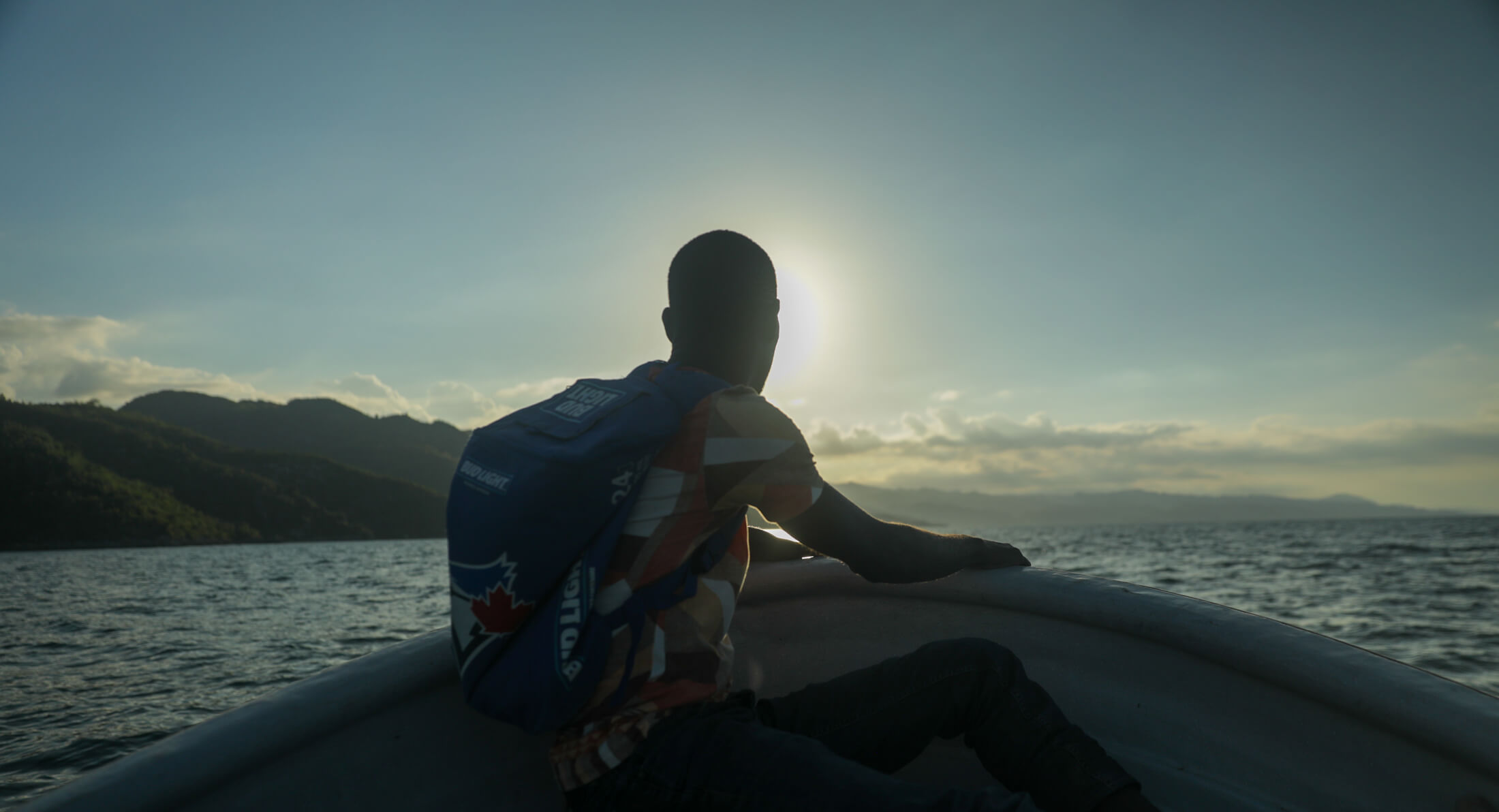 man sitting in the front of a small boat with ocean and mountains